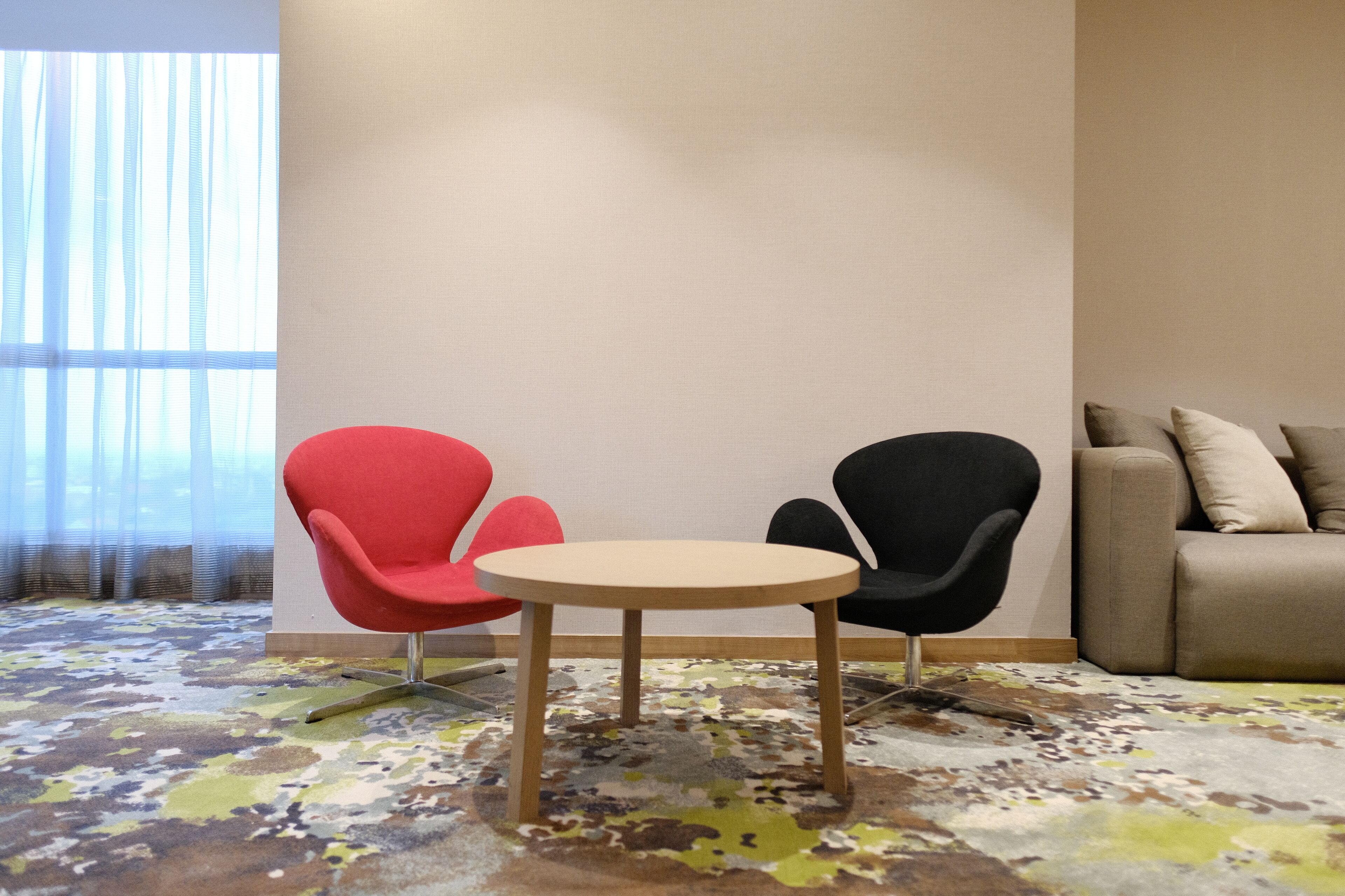 Una habitación con una mesa redonda de madera, dos sillas (una roja y una negra), un sofá color beige y una alfombra estampada.