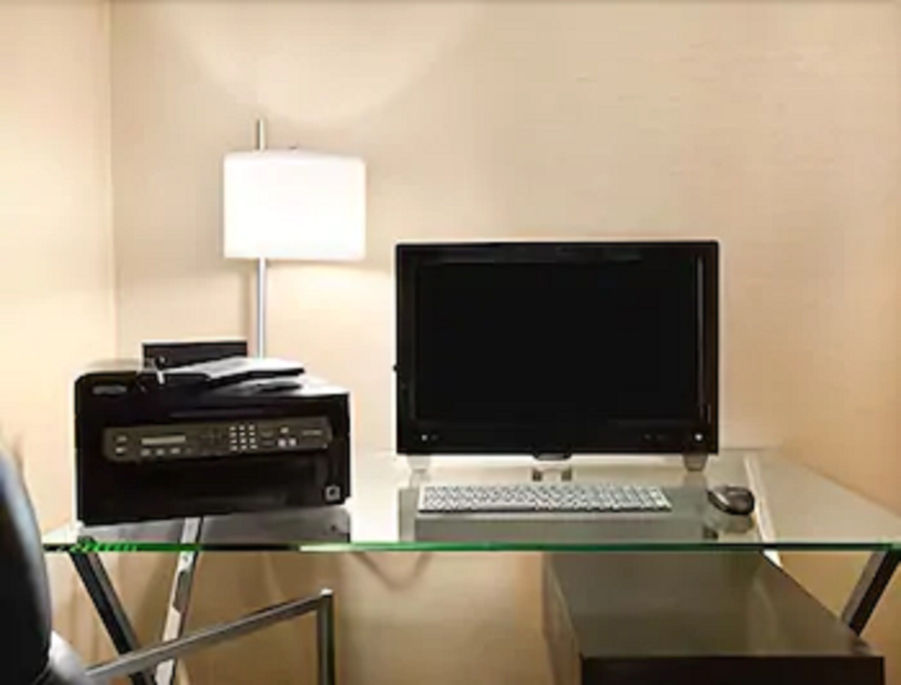 Un petit bureau avec un bureau en verre, un écran d’ordinateur, un clavier, une souris et une imprimante.