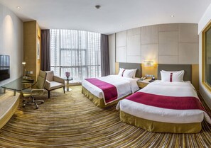 A hotel room with two beds, a desk, a chair, a TV, and a large window with curtains.
