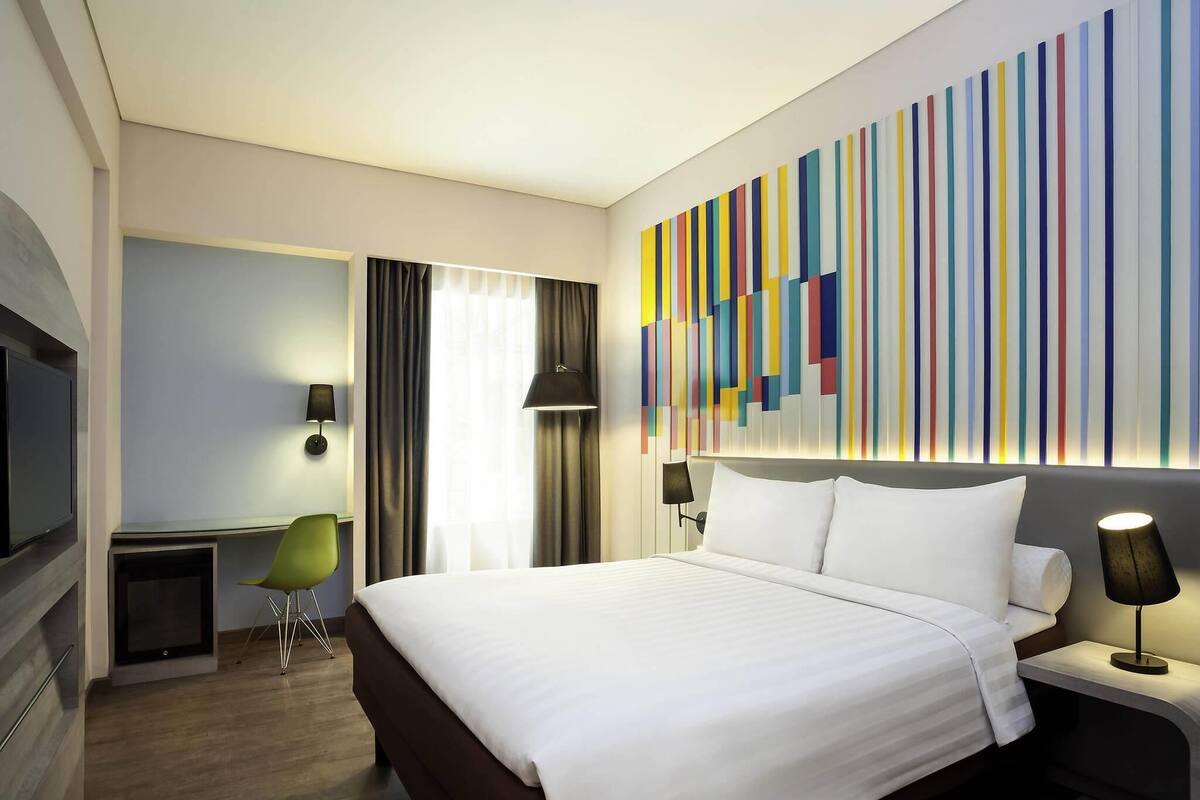 A hotel room with a large bed, a desk, a chair, a TV, and a colorful wall art piece.