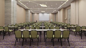 Un salón de conferencias con filas de mesas y sillas, una pantalla de proyector y suelo alfombrado.