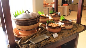 Ein Buffet mit verschiedenen Speisen, serviert in traditionellen Tongefäßen, einer Metallauflageplatte und einem Schild mit den Aufschriften 'Traditional Rice' und 'Nasi Uduk'.