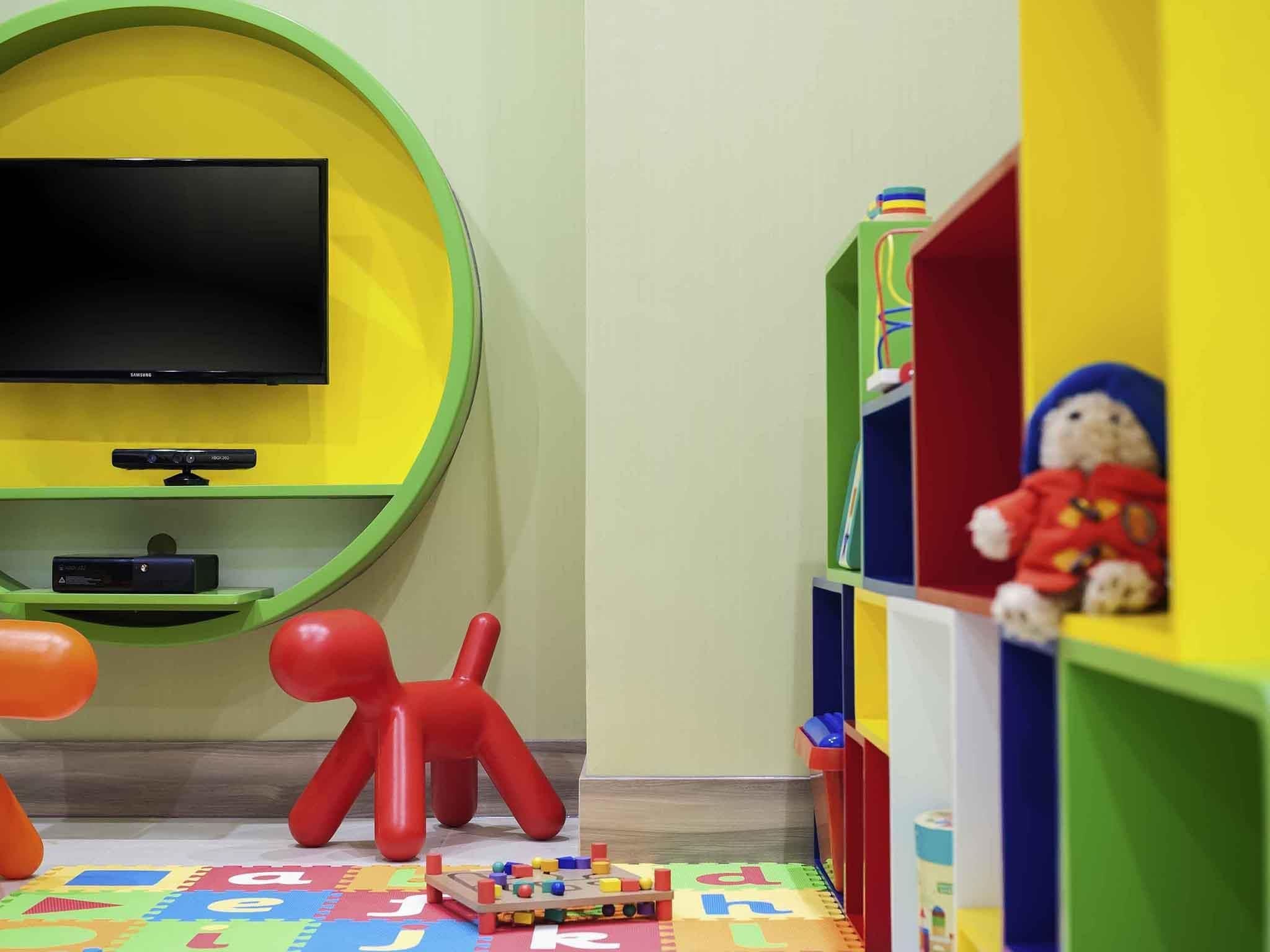 Una habitación con una televisión de pantalla plana montada en una pared verde, una figura de juguete roja y un tapete de juego con el alfabeto en el suelo.