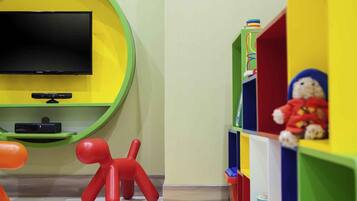 Una habitación con una televisión de pantalla plana montada en una pared verde, una figura de juguete roja y un tapete de juego con el alfabeto en el suelo.