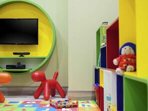 A room with a flat-screen TV mounted on a green wall, a red toy figure, and a colorful alphabet play mat on the floor. - ibis Styles Jakarta Mangga Dua Square (Jakarta)
