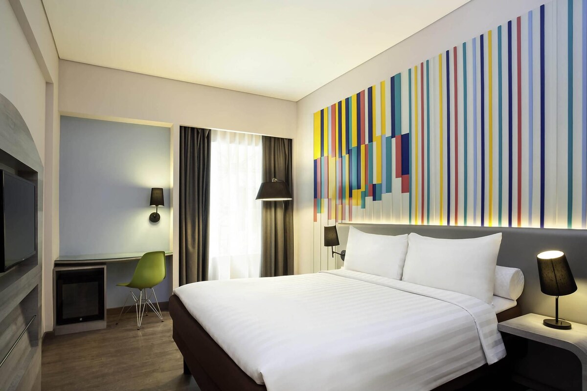 A hotel room with a large bed, a desk, a chair, a TV, and a colorful wall art piece.