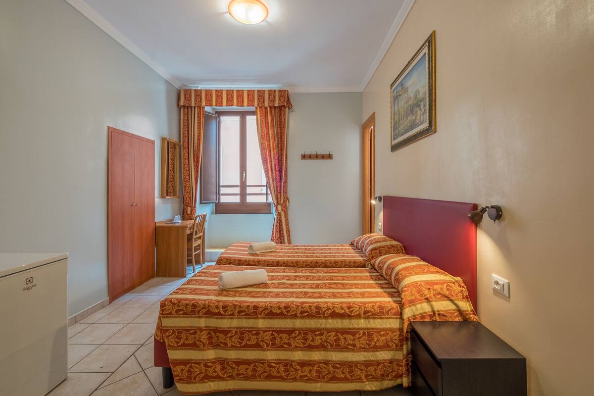 A hotel room with two beds, a desk, a chair, a wardrobe, and a window with curtains.
