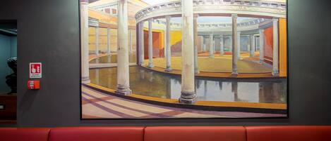 A large painting of a classical architectural scene with columns and arches, displayed on a wall above a red seating area.