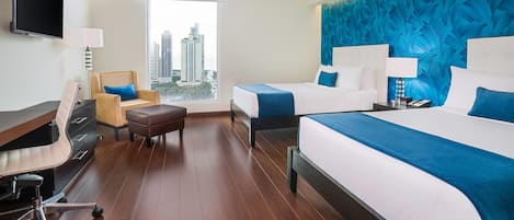 A hotel room with a large bed, two smaller beds, a desk with a chair, a TV, and a view of the city.