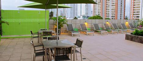 Pátio externo com mesa e cadeiras, cercado por guarda-sóis verdes.