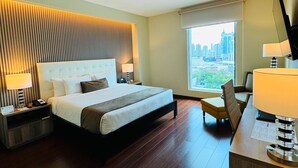 A hotel room with a large bed, a desk with a lamp, a chair, and a view of a cityscape through a window.
