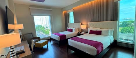 A hotel room with two beds, a desk with a lamp, a chair, and a large window with a city view.