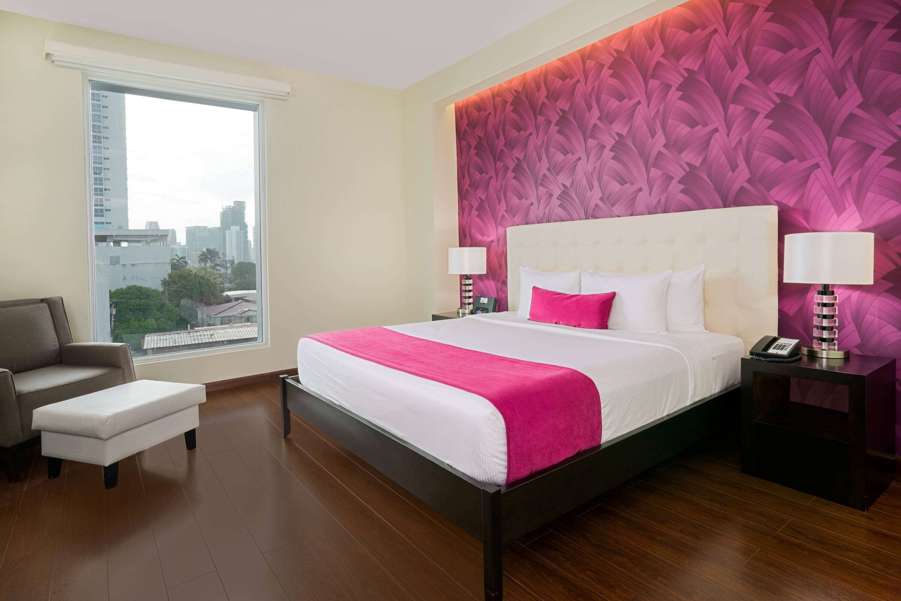 A modern hotel room with a large bed, a city view, a sofa, and a nightstand with a lamp.