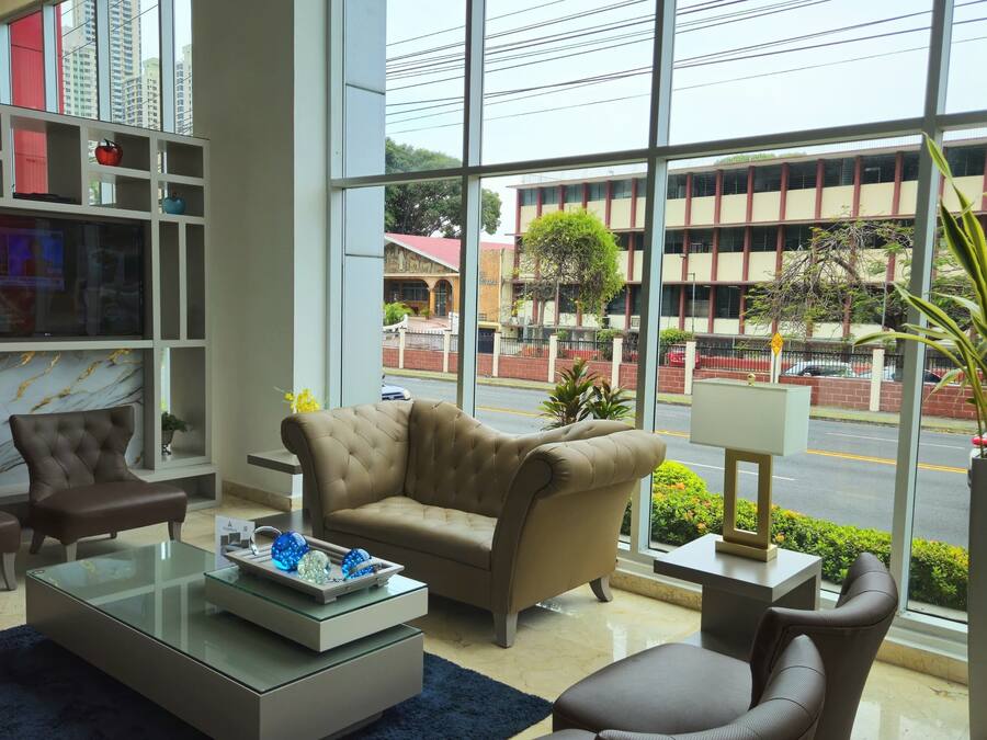 Un moderno vestíbulo de hotel con un amplio ventanal que ofrece vistas panorámicas de la ciudad, una zona de estar confortable con sofás de cuero, una chimenea de mármol y una estantería bien organizada.