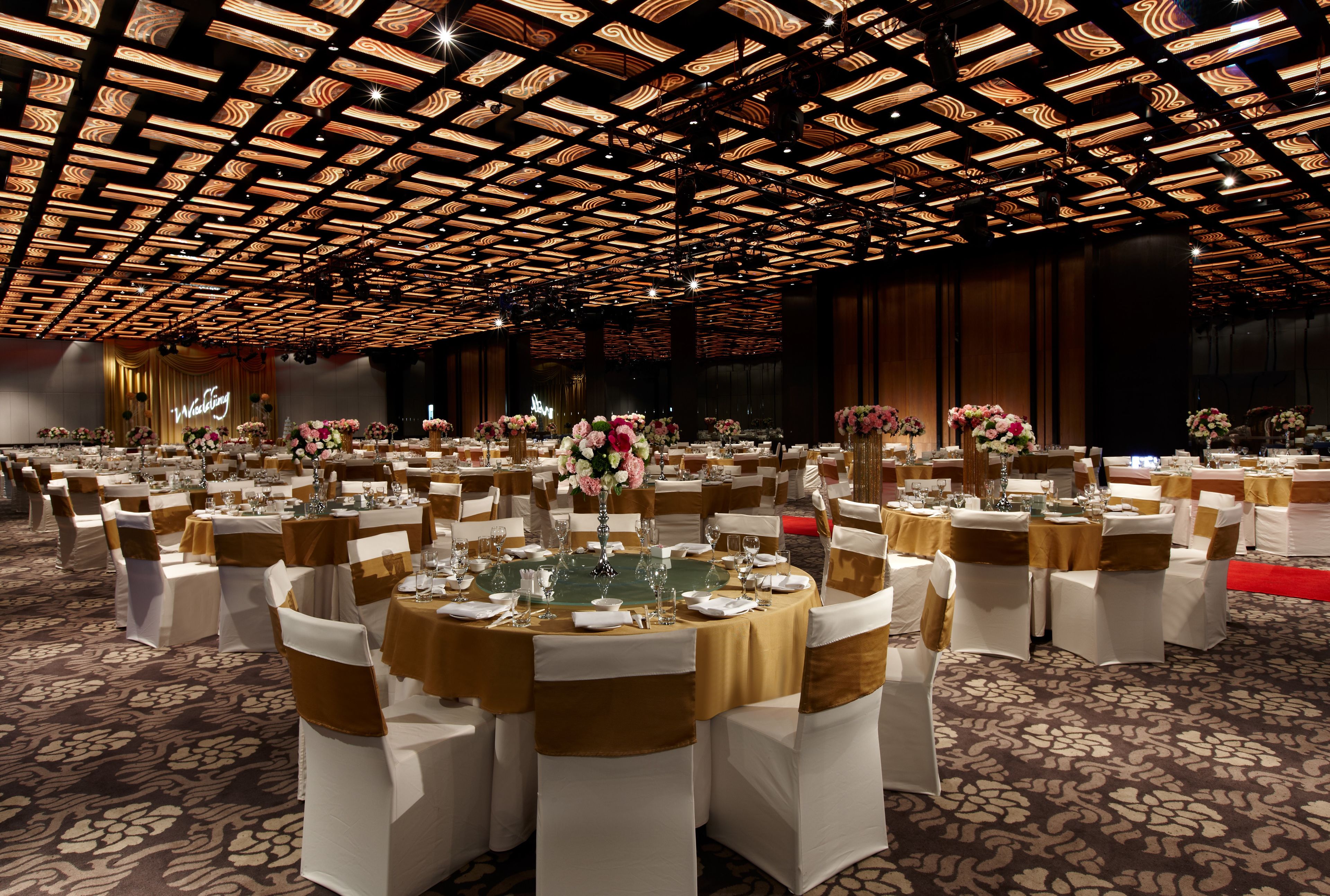 Un salón de banquetes con mesas redondas dispuestas para un evento formal, con techo estampado y suelo alfombrado.
