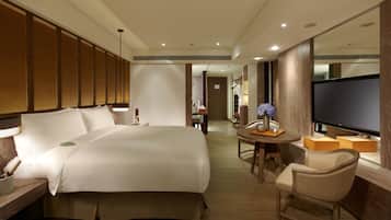 A hotel room with a large bed, a desk with a lamp, a chair, a small table with a vase, and a TV.