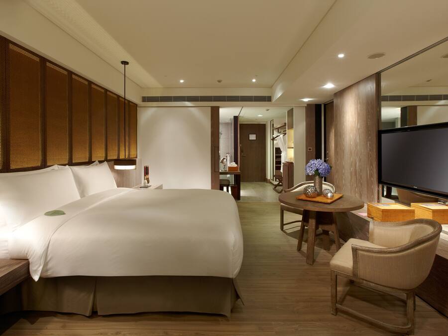 A hotel room with a large bed, a desk with a lamp, a chair, a small table with a vase, and a TV.