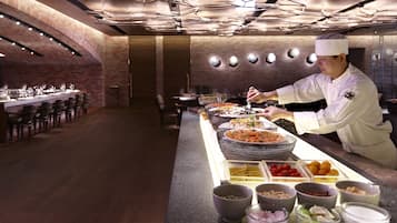 A chef preparing food in a modern kitchen with brick walls and a long counter displaying various dishes and ingredients.