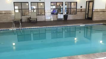 An indoor swimming pool with clear water, a tiled edge, and a seating area with chairs and a table.