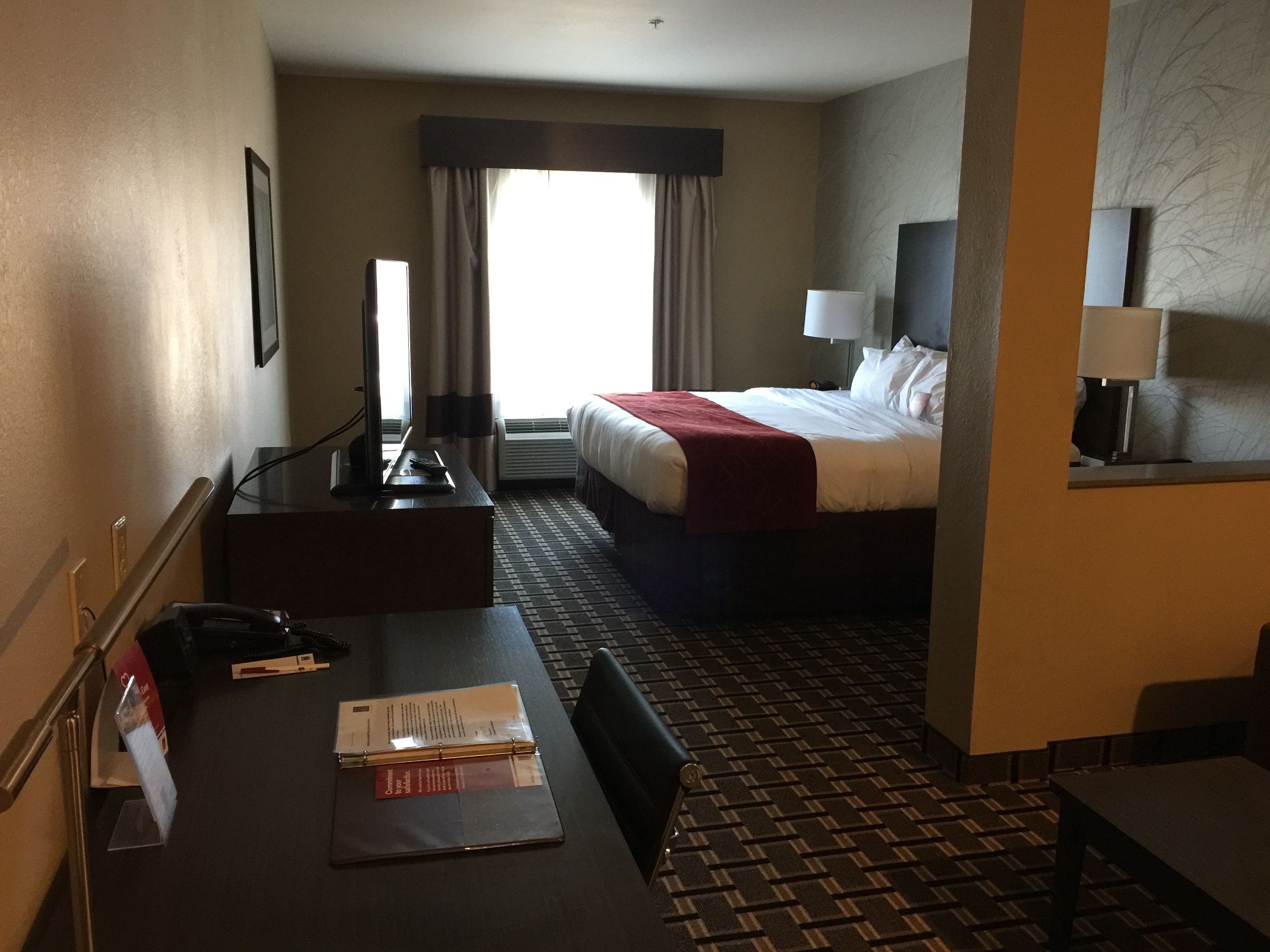 Habitación de hotel con cama, escritorio con silla, lámpara de escritorio, ventana con cortinas y alfombra estampada.
