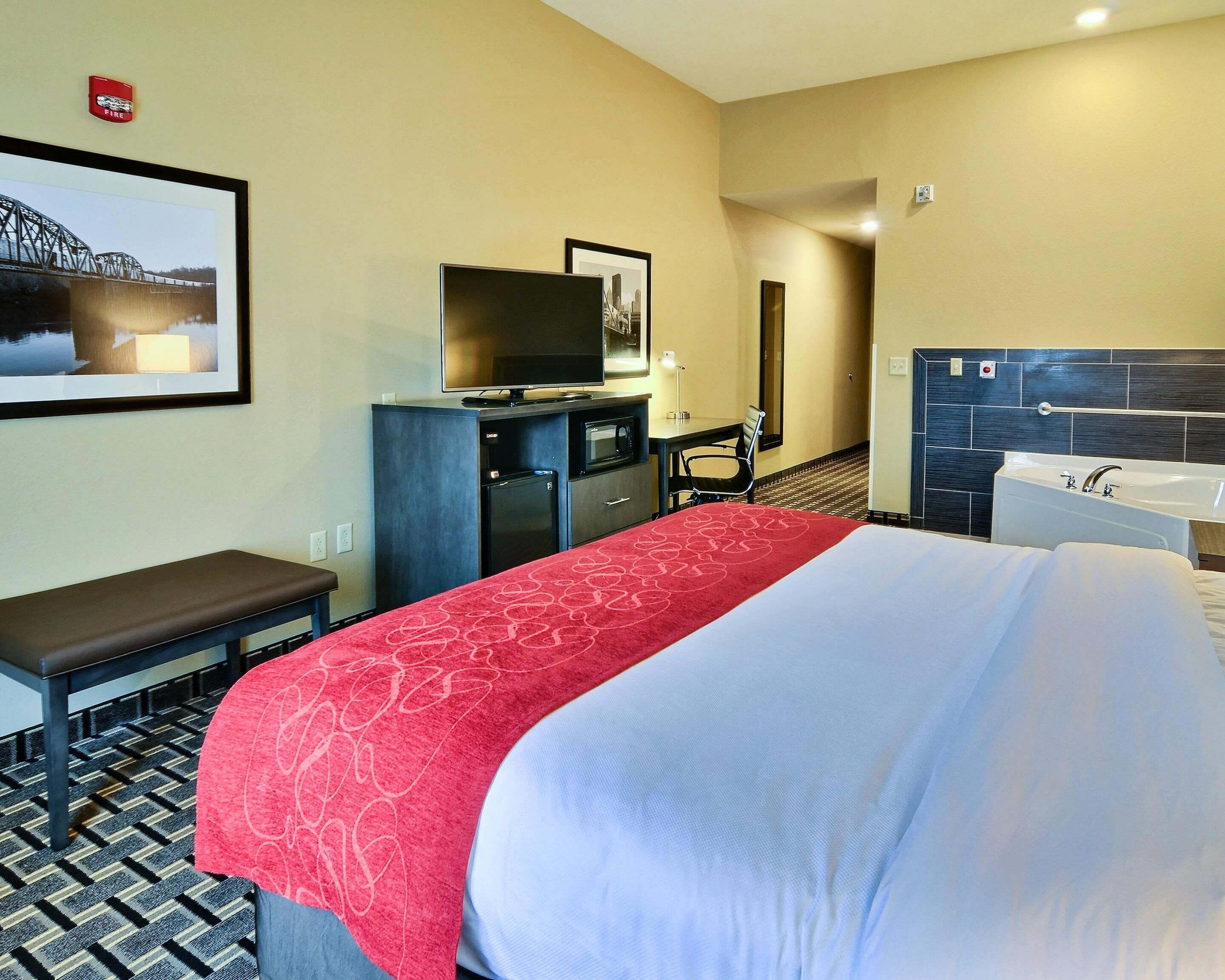 Habitación de hotel con una cama, un escritorio con silla, televisor, microondas, refrigerador, baño con bañera y detector de incendios en la pared.