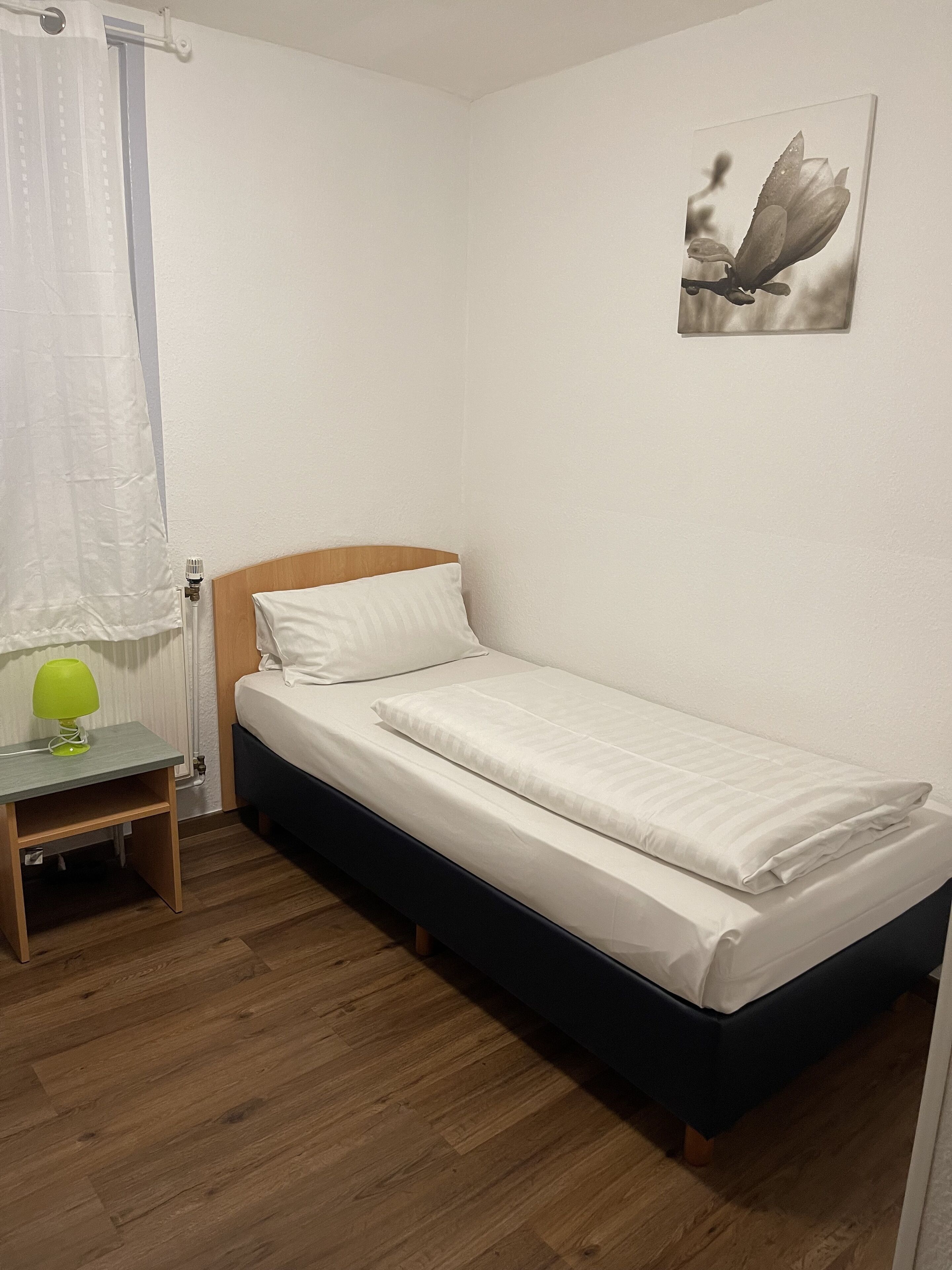 A small, neatly arranged bedroom with a single bed, a bedside table with a lamp, and a wall-mounted picture.