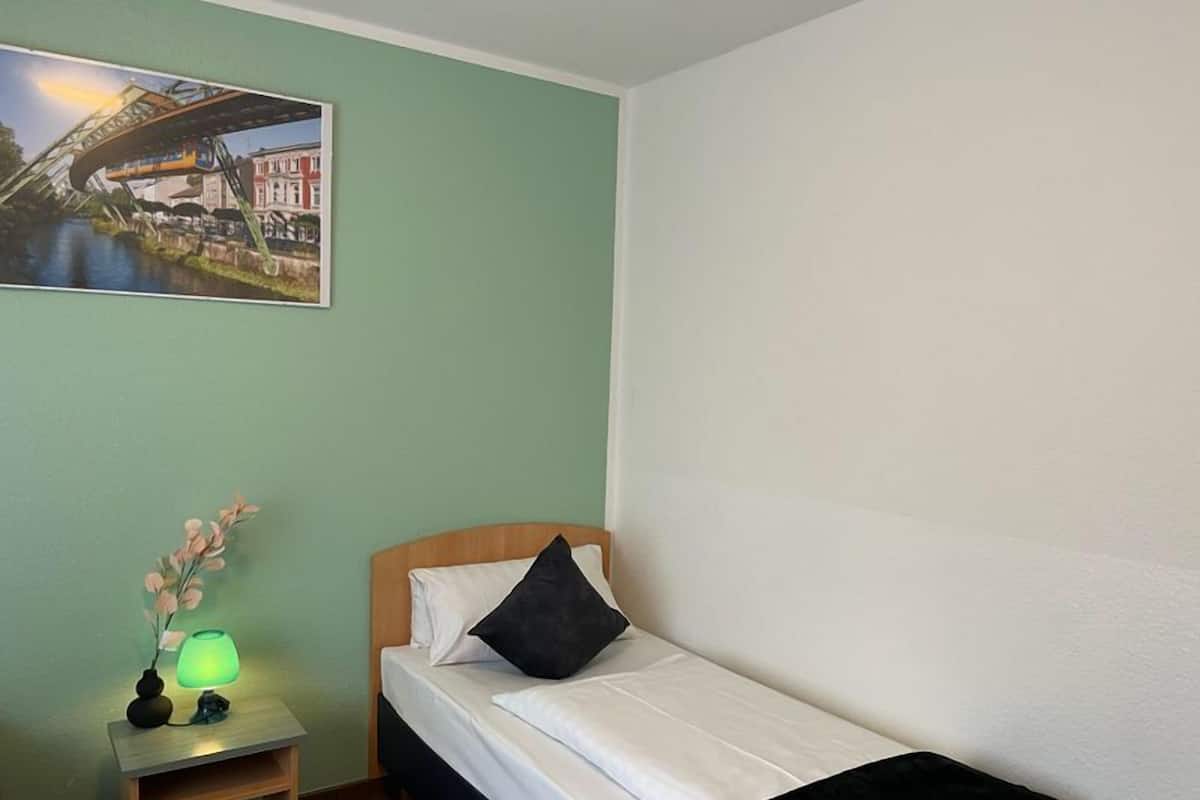 Une petite chambre à coucher, bien rangée, avec un lit simple, une table de chevet avec une lampe et un tableau encadré au mur.