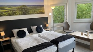 Une chambre d’hôtel avec deux lits, un bureau, une chaise, une lampe et une fenêtre donnant sur un paysage verdoyant.