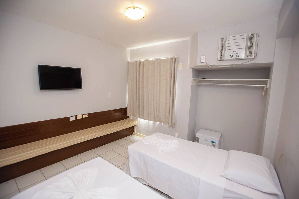 A hotel room with a bed, a bench, a television, a window with curtains, and a wall-mounted air conditioning unit.