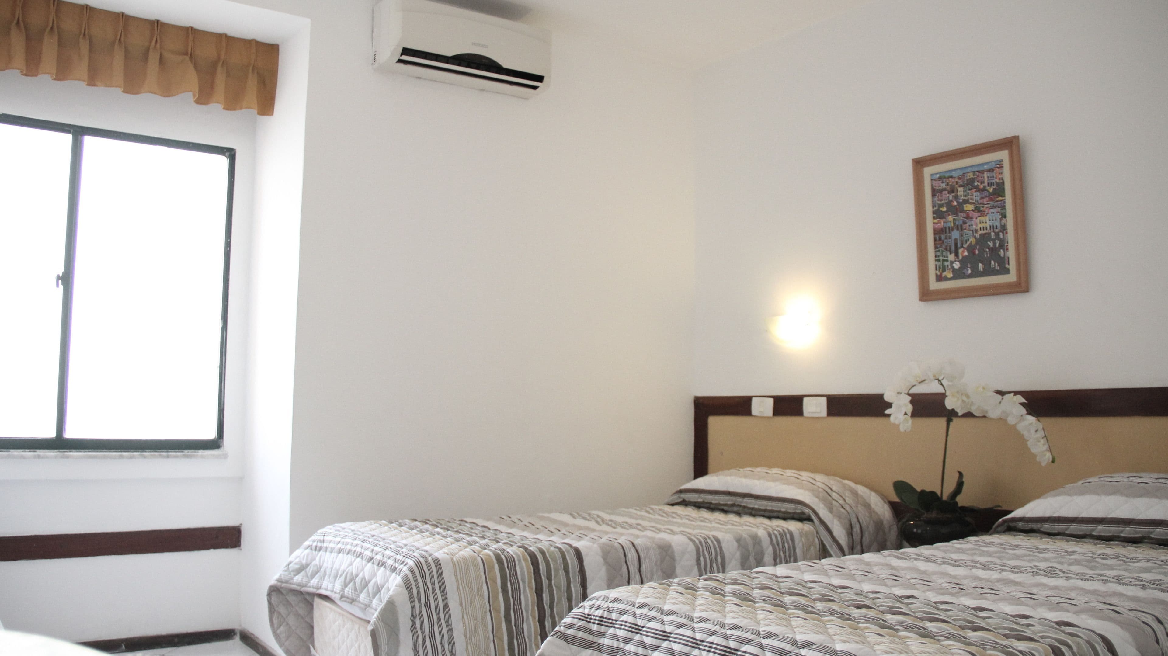 Habitación de hotel con dos camas, ventana con cortinilla marrón, unidad de aire acondicionado, luz empotrada en la pared y un cuadro enmarcado.