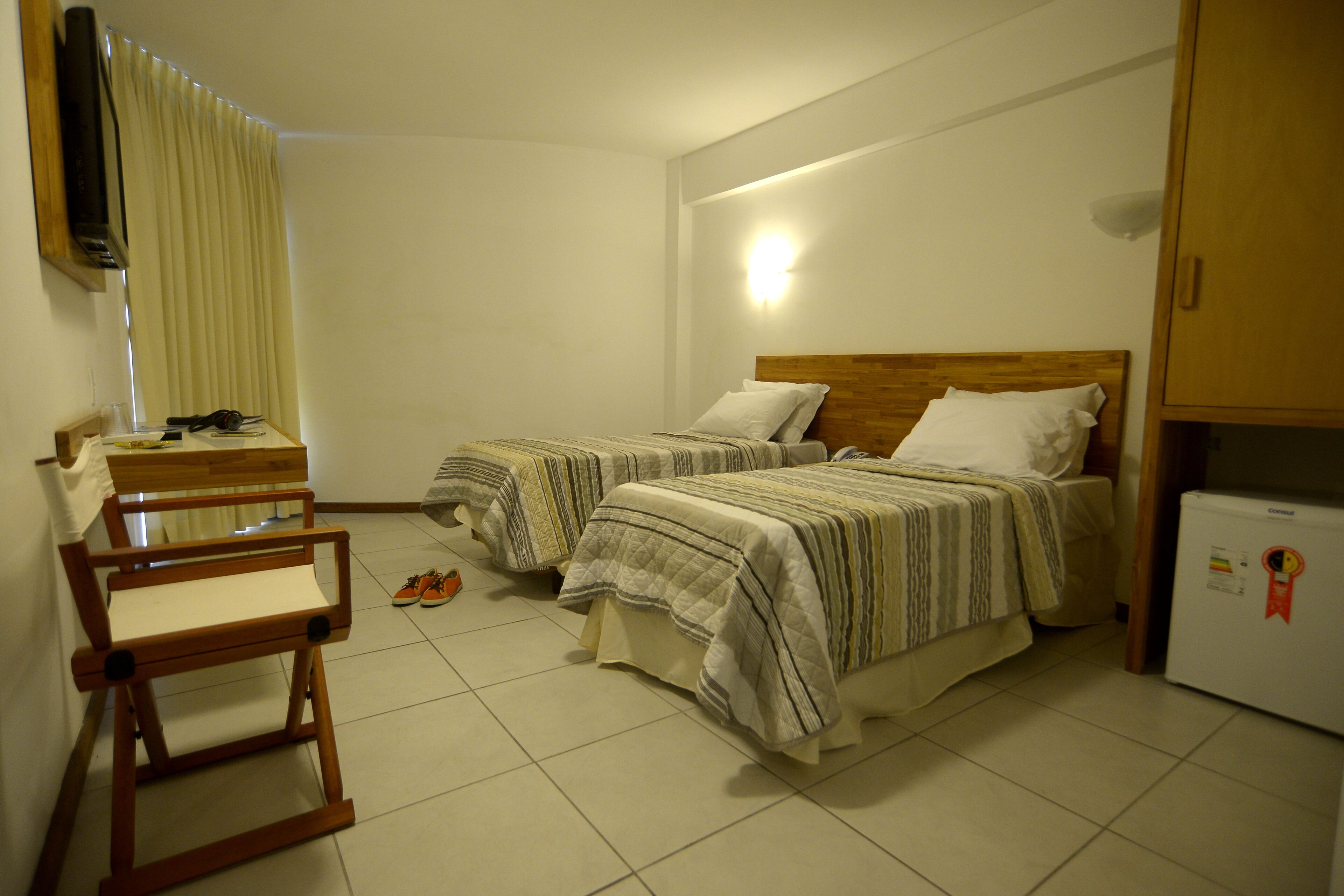 A bedroom with two beds, a wooden chair, a small table, a television, and a refrigerator.