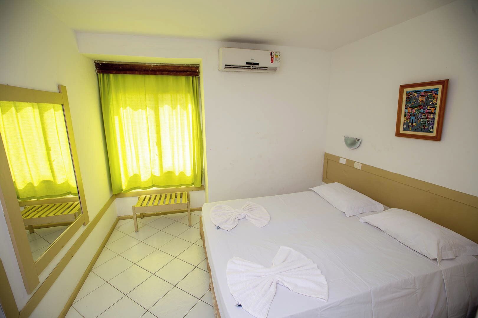 A hotel room with a bed, a bench, a window with curtains, an air conditioning unit, and a wall-mounted light fixture.