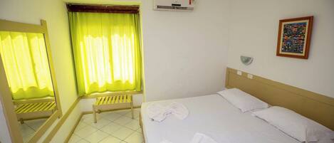 A hotel room with a bed, a bench, a window with curtains, an air conditioning unit, and a wall-mounted light fixture.