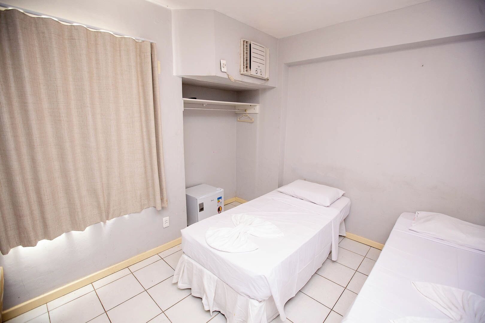 A small, clean room with a single bed, a bedside table, a heater, and a window with curtains.