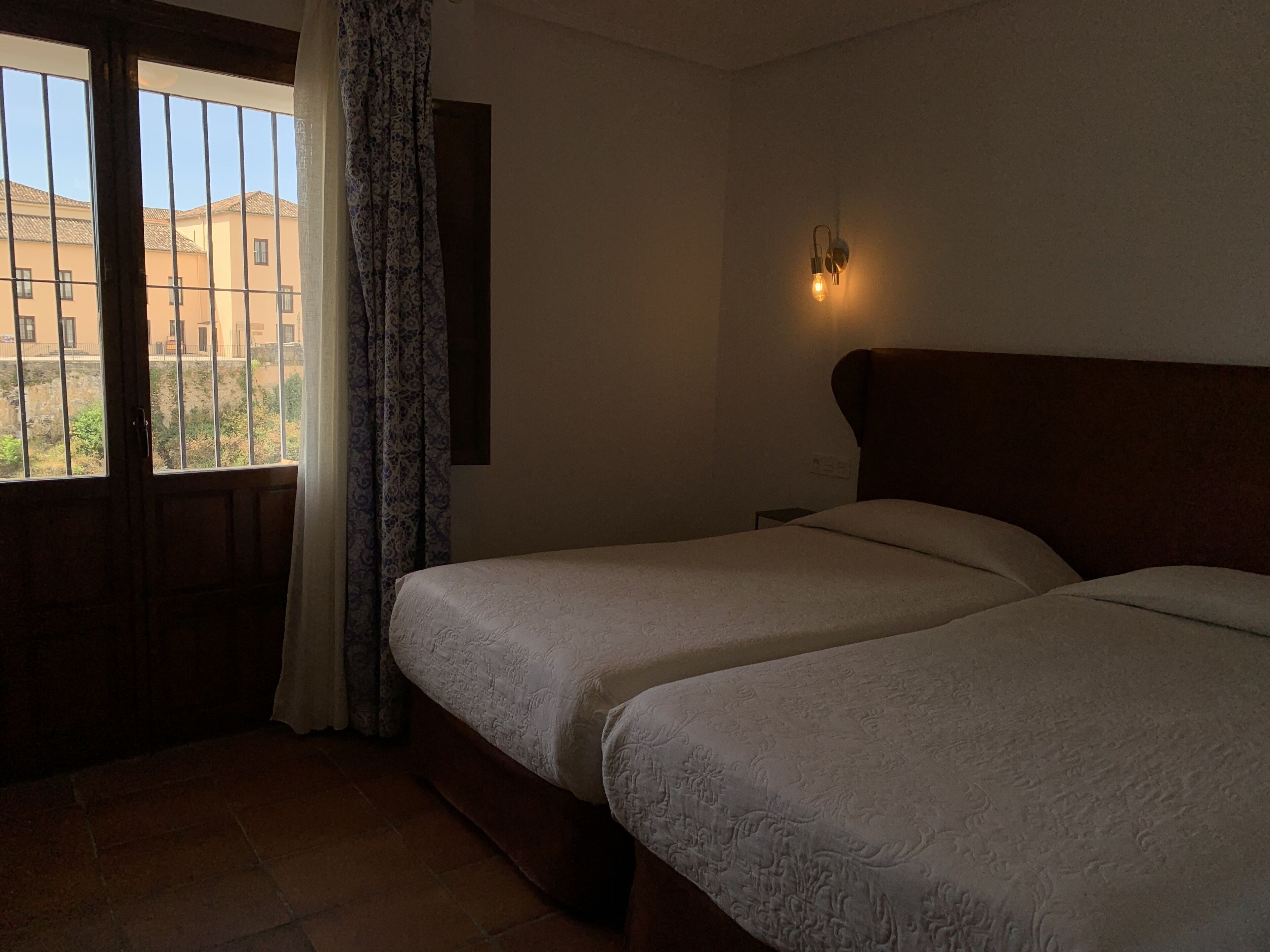 A hotel room with two beds, a window with curtains, and a wall-mounted light fixture.