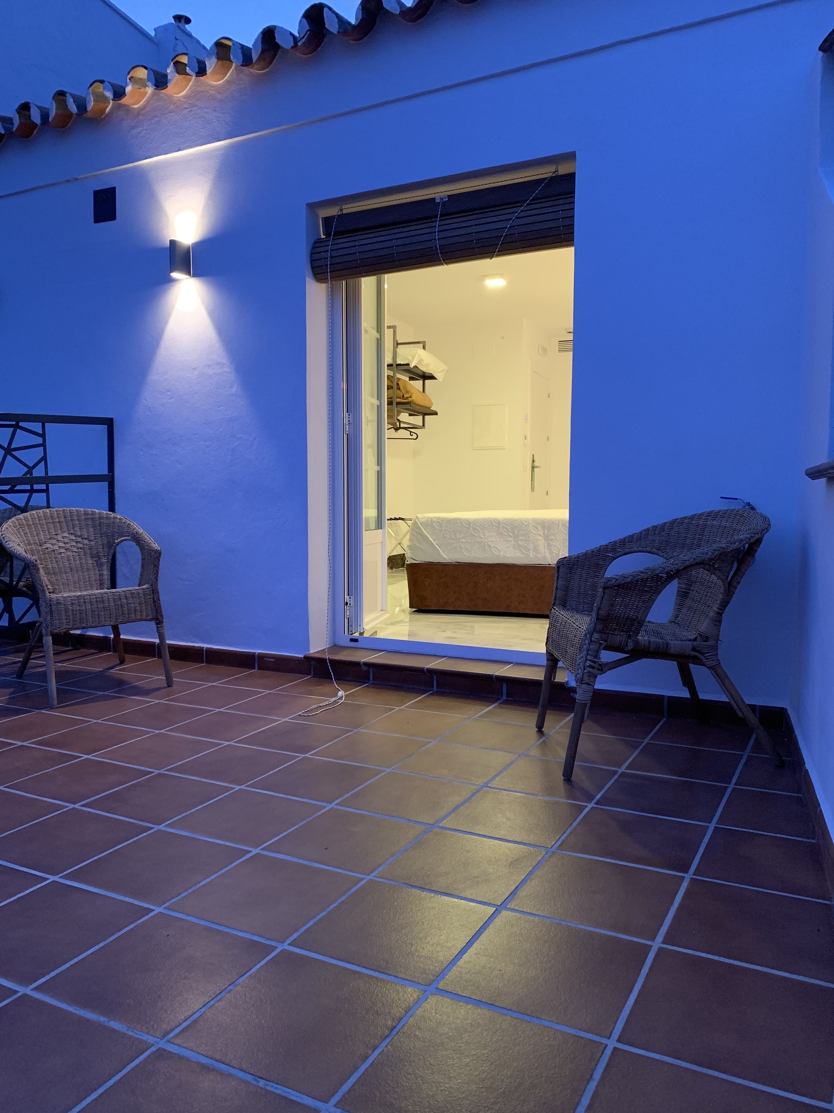 A room with a tiled floor, a bed, a chair, and a wall-mounted shelf.