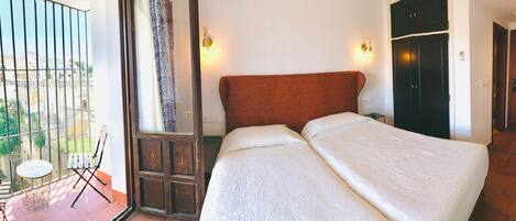 Un dormitorio con cama, ventana con vista a edificios, puerta y una pequeña mesa con silla.