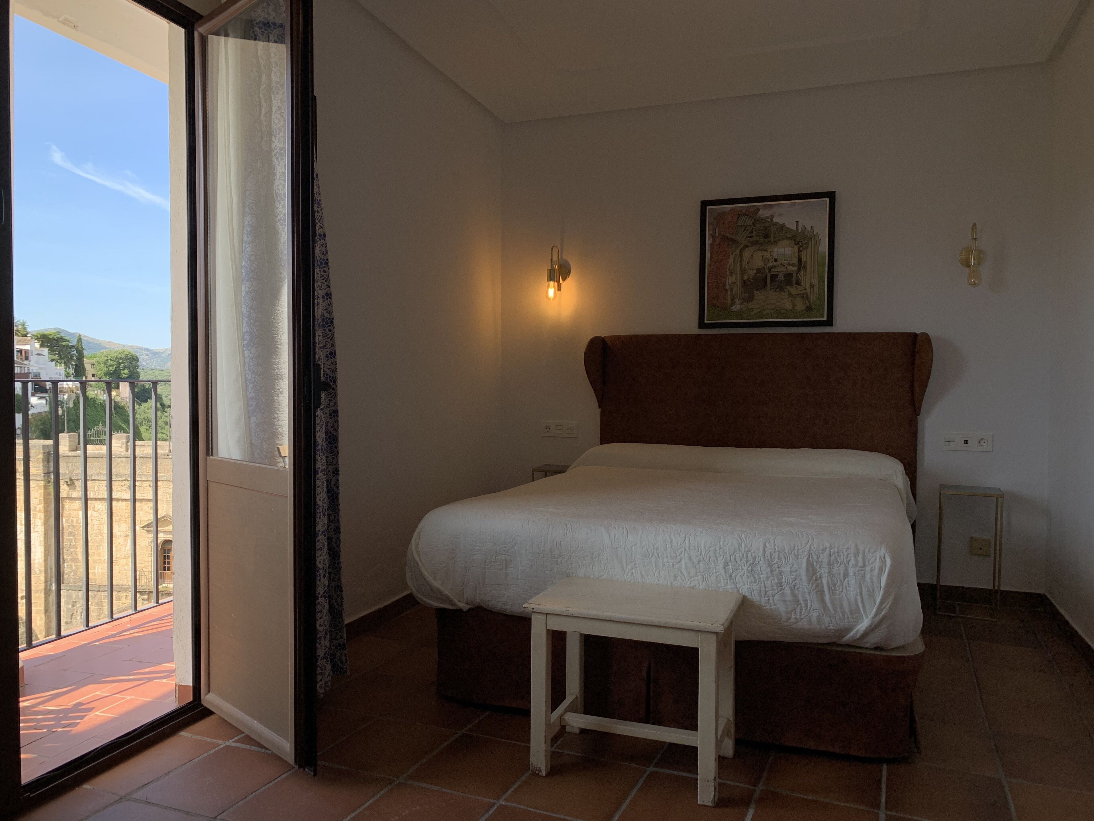 Een slaapkamer met een bed, een klein nachtkastje, een schilderij aan de muur en uitzicht door een raam.