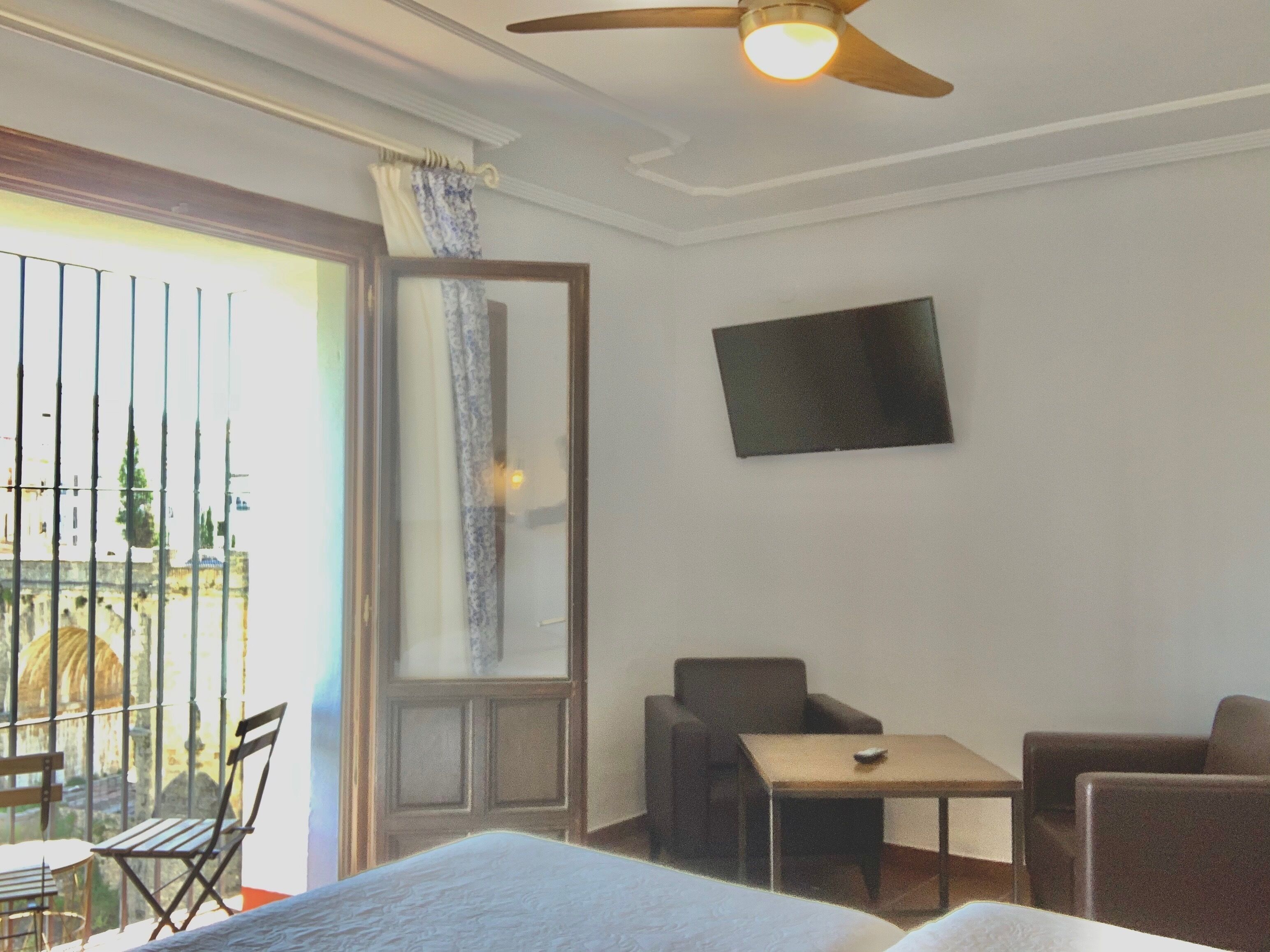 A hotel room with a bed, a desk, two chairs, a TV on the wall, and a balcony with a view.