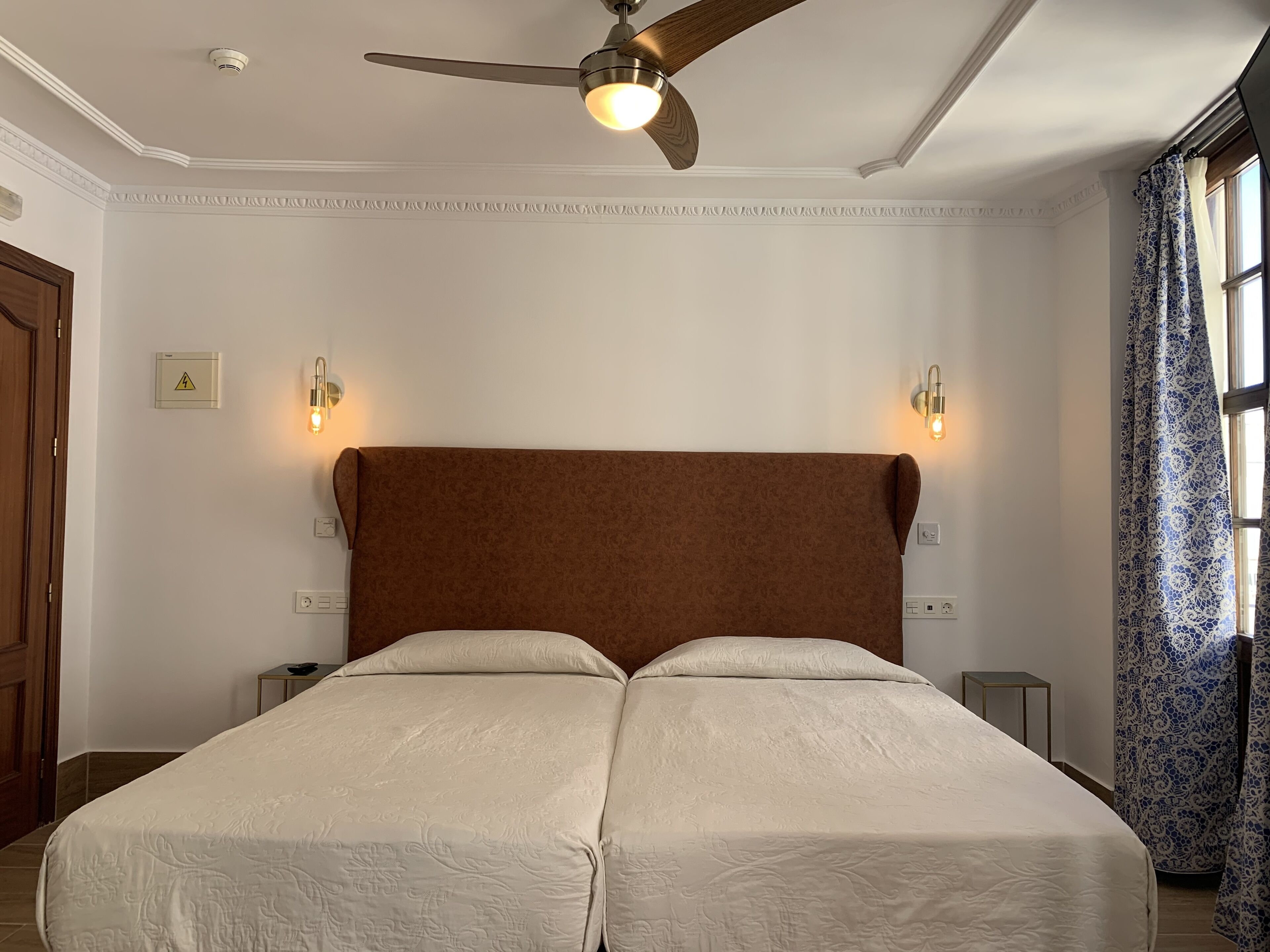 A double bed with a brown headboard, two bedside tables, a ceiling fan, and a window with patterned curtains.