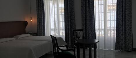 A hotel room with a bed, two chairs, a table, and large windows with patterned curtains.