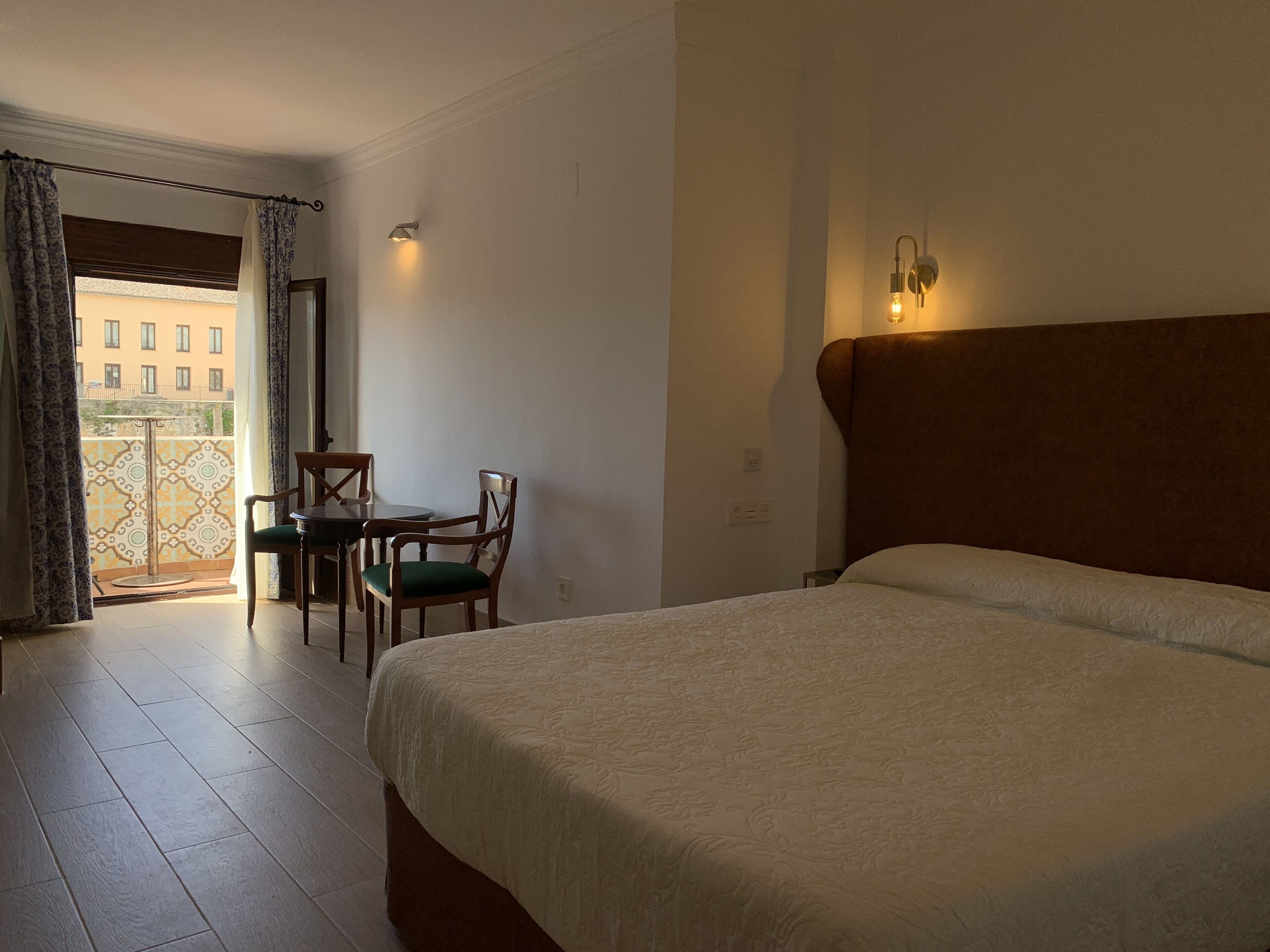 A hotel room with a bed, a small table with chairs, a balcony with patterned curtains, and a wall-mounted light fixture.