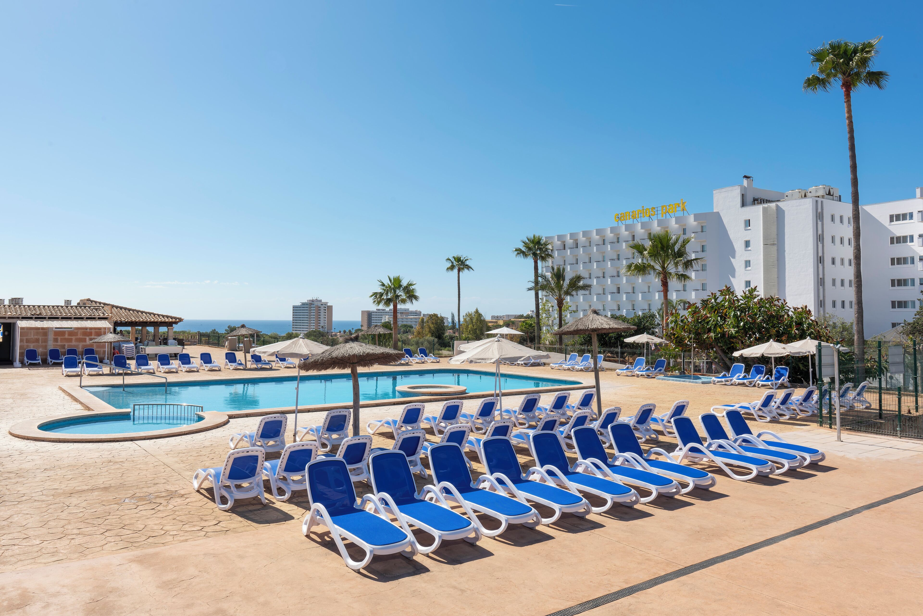 A poolside area with blue lounge chairs and umbrellas, a clear sky, and a building with a sign that reads "Camino de Santiago park".