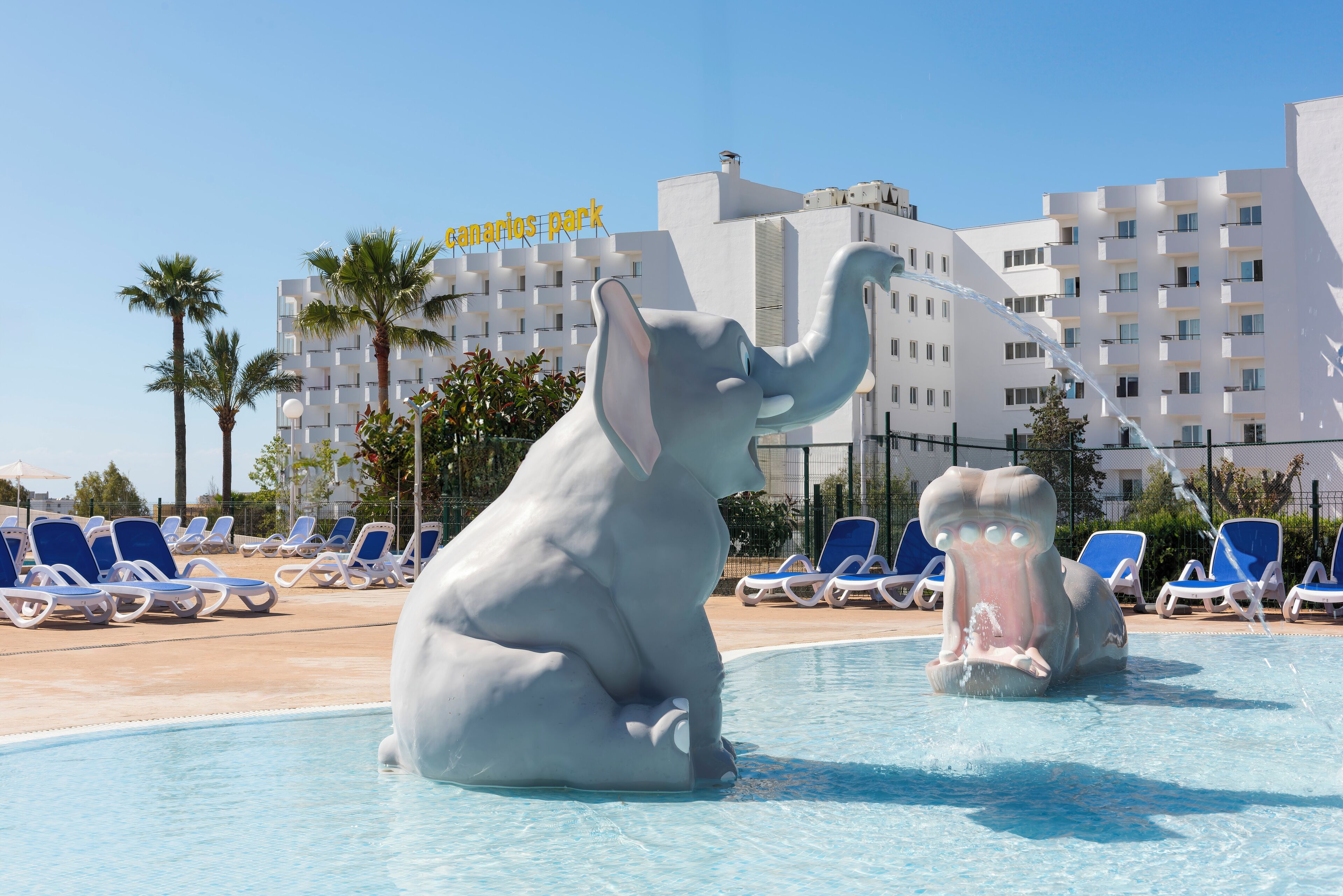 Ein Poolbereich mit zwei Elefantenstatuen, blauem Himmel und einem Gebäude mit Balkonen im Hintergrund.