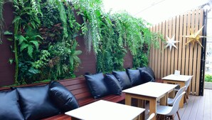 Uma área de pátio com bancos e mesas de madeira, rodeada por vegetação exuberante e luzes decorativas em forma de estrela.