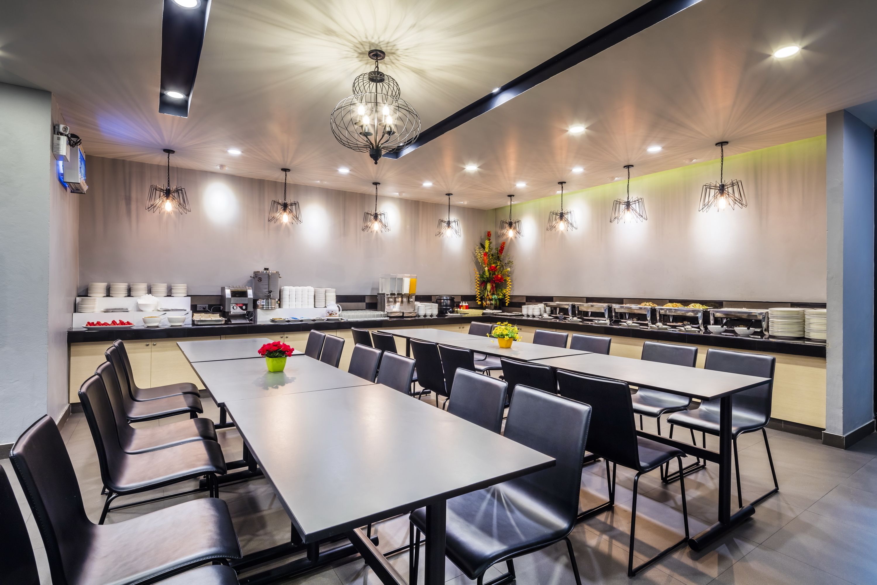 Un comedor moderno con mesas largas y sillas, una lámpara decorativa en el centro y un mostrador de buffet con alimentos.