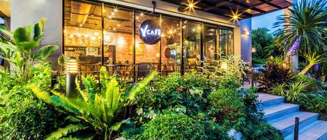 Un café moderne doté d’une terrasse, entouré d’une végétation luxuriante et d’un jardin bien entretenu.
