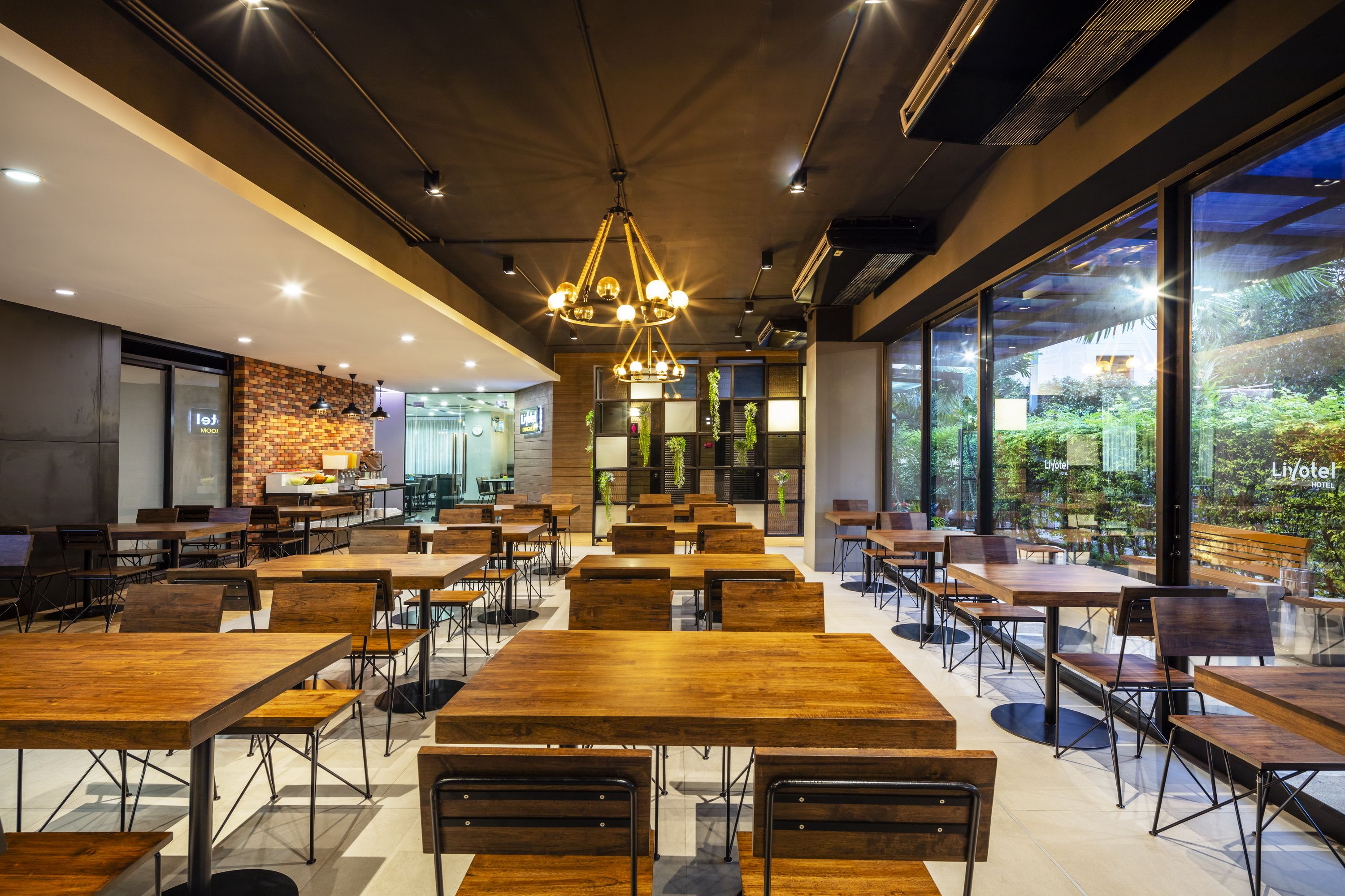 A spacious indoor dining area with wooden tables and chairs, a modern chandelier, and large windows offering views of greenery.