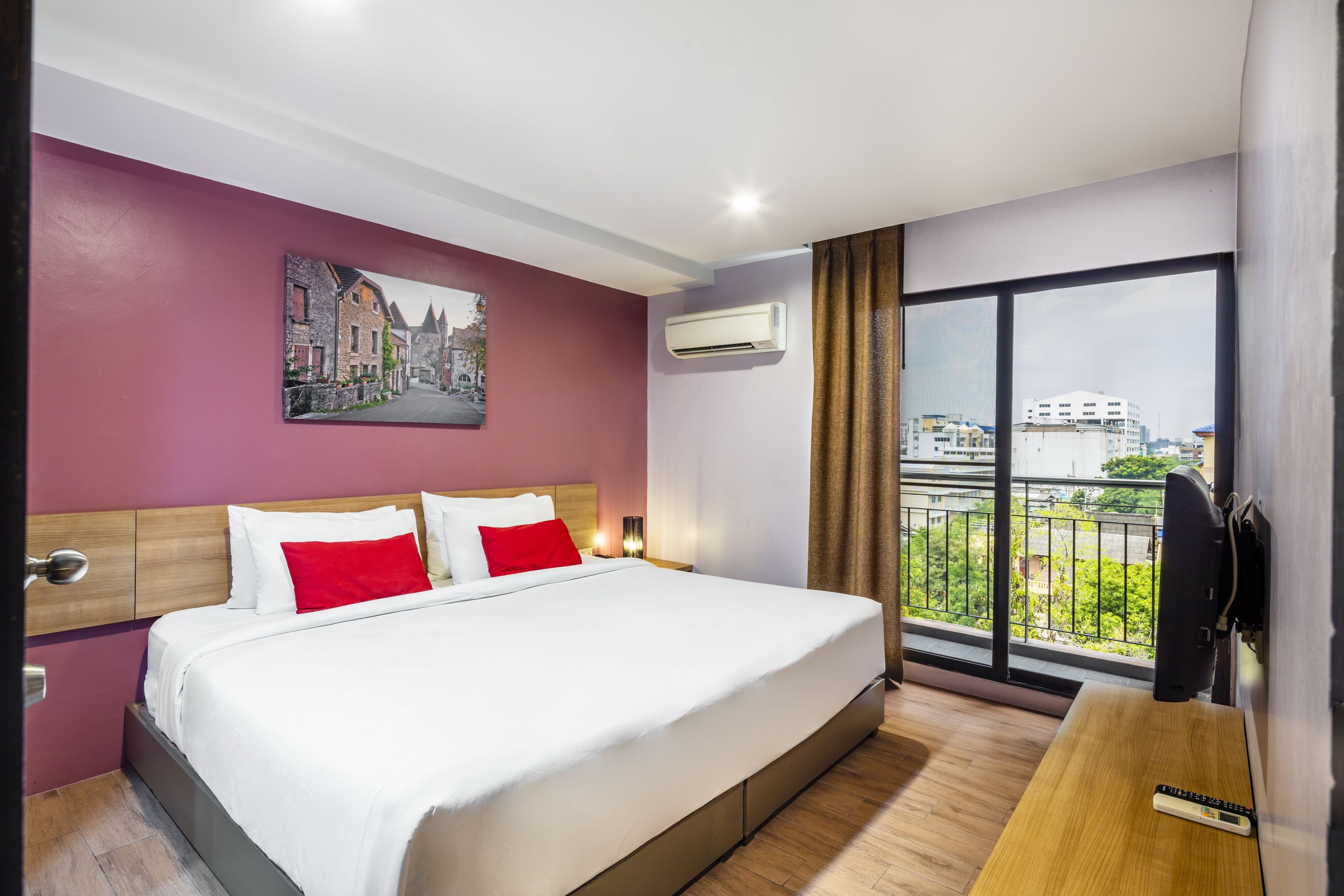 A hotel room with a large bed, a painting of a street scene, an air conditioning unit, a balcony with a view of buildings, and a desk with a remote control.