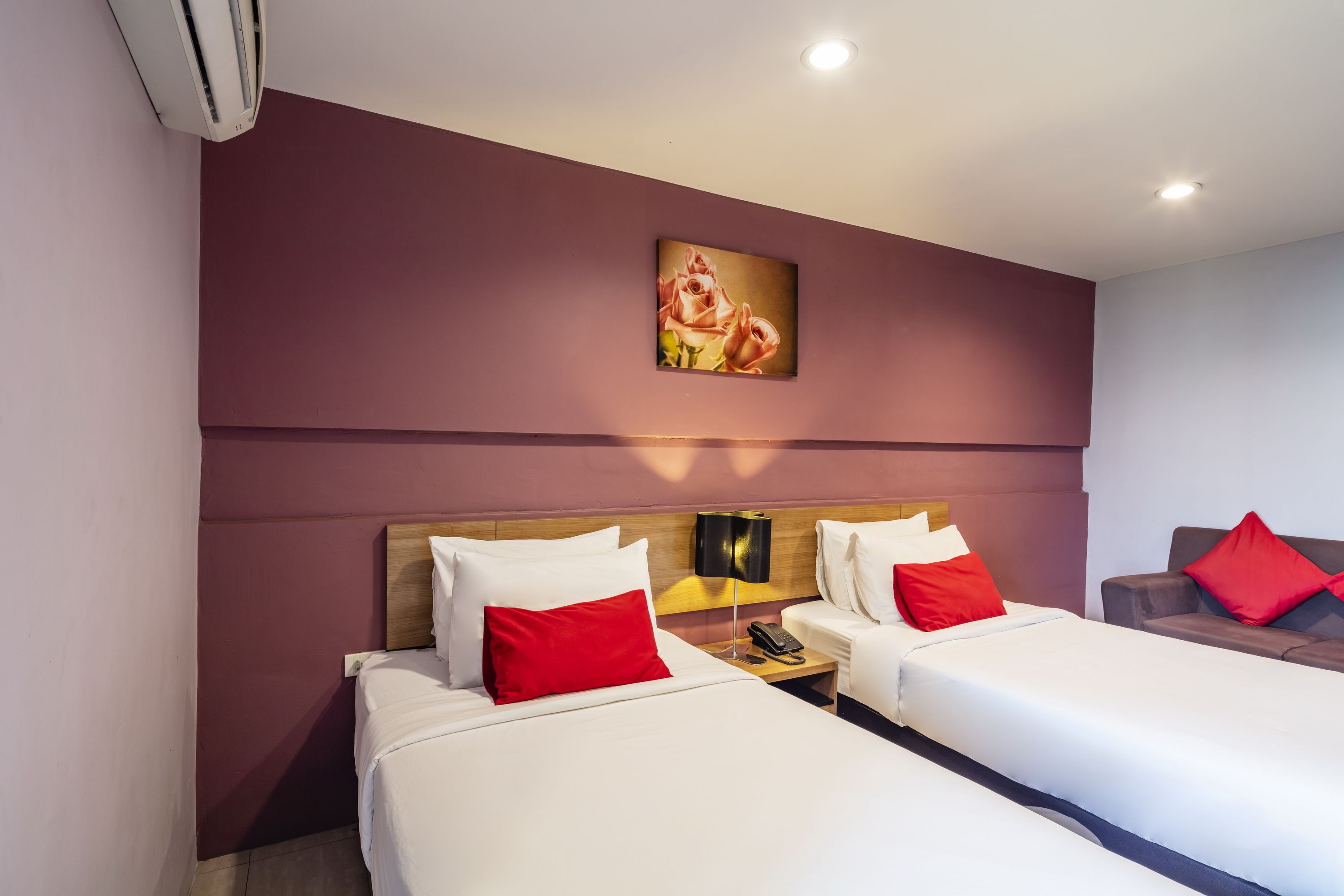 A hotel room with two beds, each with white bedding and red pillows, a wooden headboard, a nightstand with a phone, and a painting of flowers on the wall.